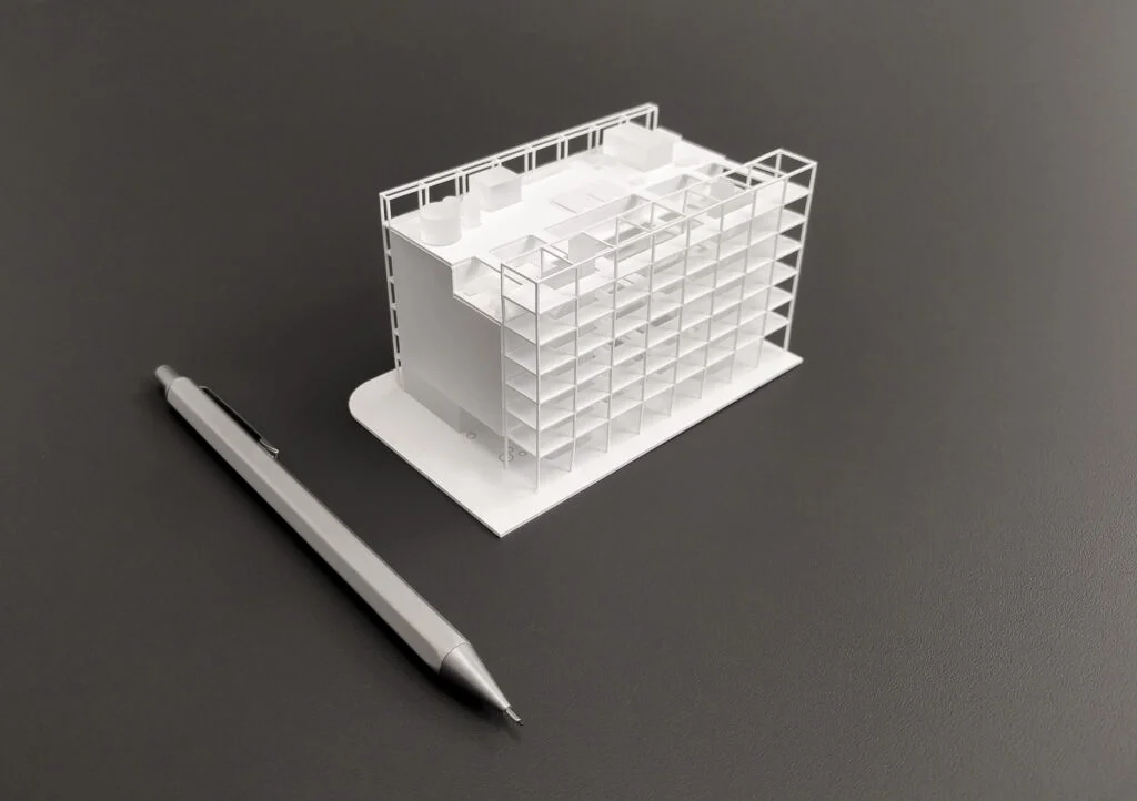 Architekturmodell – einfaches weißes Arbeitsmodell im Maßstab 1:500 mit klarer Fassadenstruktur und hohem Detailgrad, Stift als Größenvergleich.
