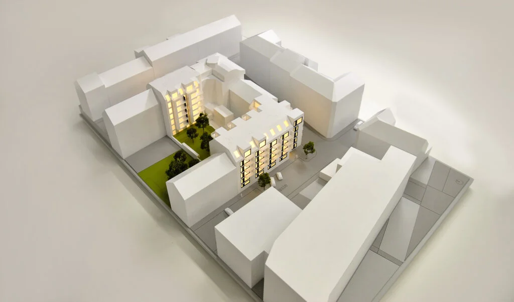 Architekturmodell Wohnblock – Immobilienmodell mit beleuchtetem Innenhof, Umgebungsbebauung und Grünflächen.