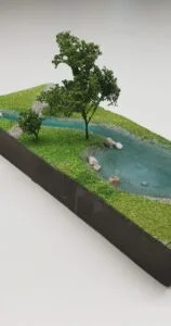 Bemusterungsmodell Flusslauf mit Bäumen, Wasserfläche, Steinen und grüner Landschaftsgestaltung.