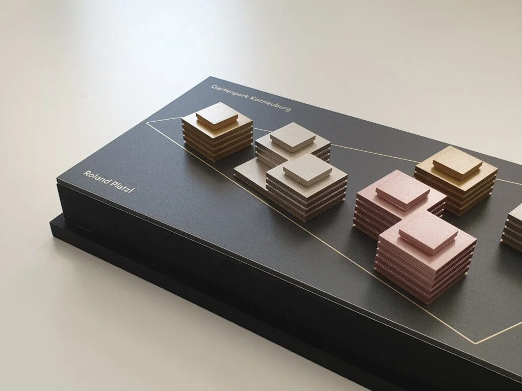 Metallmodell – abstrahierte Gebäudegruppen mit goldenen, silbernen und roséfarbenen Kuben auf schwarzem Sockel.