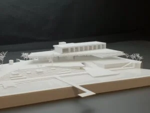 Weißes 3D-Druck Architekturmodell mit Pavillonbau, Terrassenflächen und Stufenanlage, ergänzt durch Bäume, auf dunklem Hintergrund.