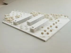 Weißes 3D-Druck Modell mit zwei langen Gebäuden, umgeben von vielen Bäumen, auf heller Grundplatte präsentiert.