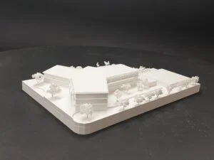 Weißes 3D-Druck Modell eines Gebäudekomplexes mit mehreren Baukörpern und Bäumen, auf dunklem Hintergrund präsentiert.