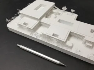 Weißes 3D-Druck Modell mit mehreren kubischen Baukörpern und Innenhöfen, platziert auf dunklem Hintergrund neben Kugelschreiber.