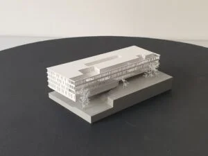 Weißes 3D-Druck Modell eines länglichen Gebäudes mit Fassadendetails und Bäumen, auf rechteckiger Grundplatte präsentiert.