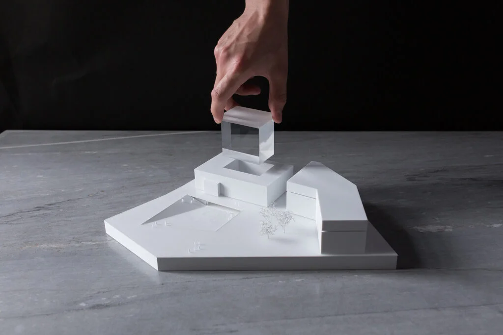 Architektur-Wettbewerbsmodell mit abnehmbarem transparentem Volumen, von einer Hand angehoben, auf minimalistischem weißen Sockel.