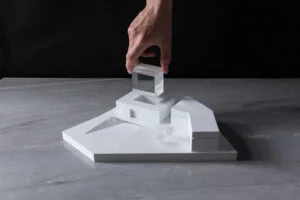 Architektur-Wettbewerbsmodell mit abnehmbarem transparentem Volumen, von einer Hand angehoben, auf minimalistischem weißen Sockel.