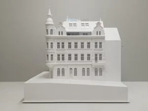 Weißes Architekturmodell eines historischen Gebäudes mit verzierter Fassade, Erkern und Turm auf einem geneigten Sockel im Maßstab 1:100.
