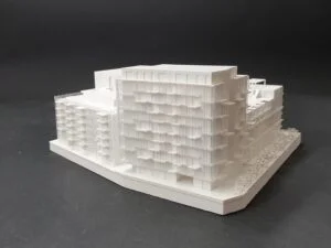 Wettbewerbsmodell eines mehrgeschossigen Wohngebäudes mit strukturierter Fassade und Bäumen auf schwarzem Hintergrund im Maßstab 1:500.