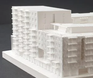 Weißes Wettbewerbsmodell mit blockartiger Gebäudestruktur, gestaffelten Volumen und Innenhof auf schwarzem Hintergrund dargestellt.