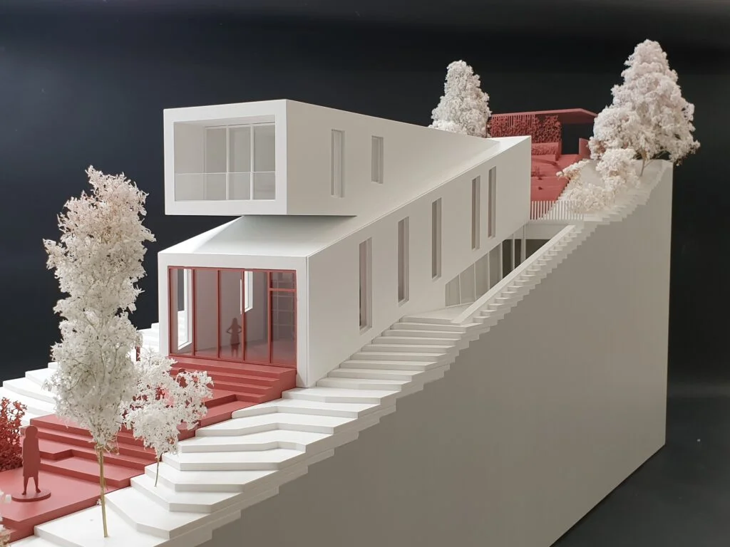 Weißes Architekturmodell eines Wohnhauses auf terrassierter Hanglage mit roten Außenflächen und Bäumen im Maßstab.