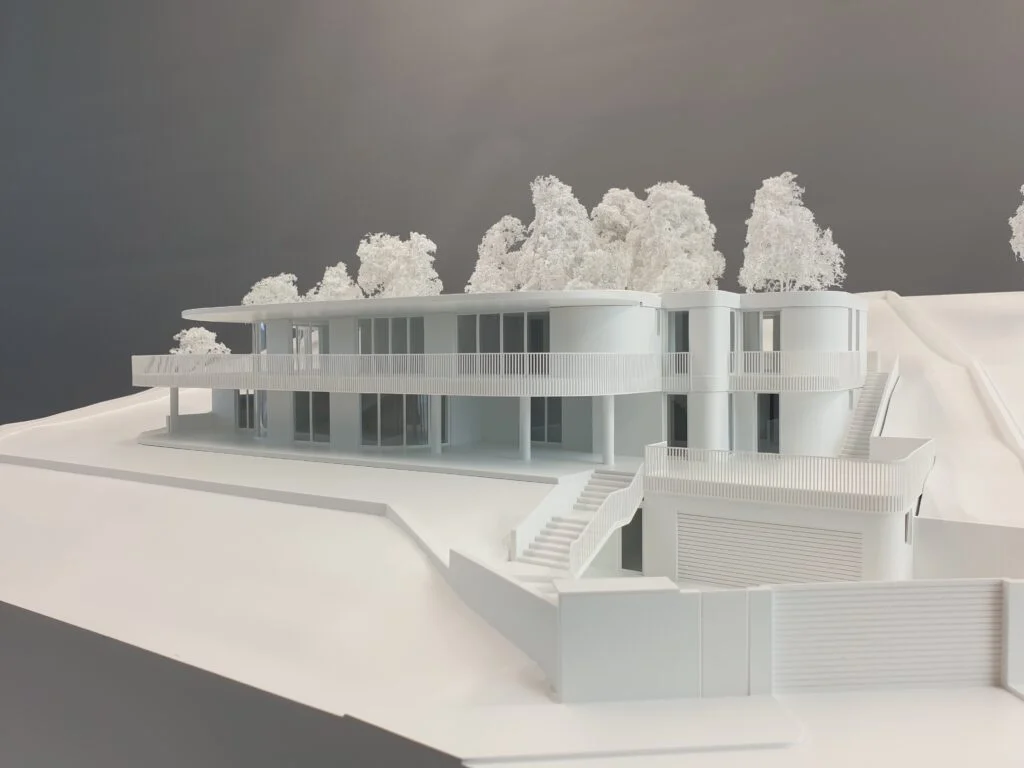Weißmodell einer terrassierten Villa mit modernen Rundformen und umlaufenden Balkonen in Hanglage, umgeben von weißen Modellbäumen.