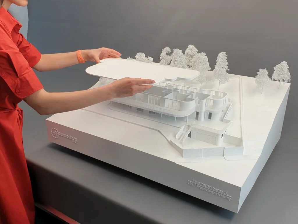 Weißes Architektur-Weißmodell mit abnehmbarer Geschossplatte, gehalten von einer Person, mit umliegendem Gelände und Bäumen.
