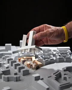 Hand platziert ein weißes Volumen auf einem städtebaulichen Ausstellungsmodell mit grauen Gebäuden und Holzbausteinen.