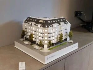 Beleuchtetes Architekturmodell eines modernen Wohngebäudes mit Balkonen, Begrünung und detaillierter Straßensituation.