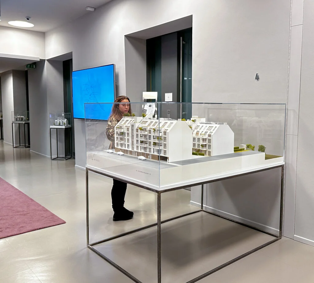 Architekturmodell in Glasvitrine im modernen Ausstellungsraum, Präsentation eines Wohnbauprojekts mit Besucherinteraktion.
