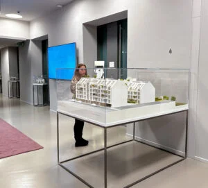 Architekturmodell in Glasvitrine im modernen Ausstellungsraum, Präsentation eines Wohnbauprojekts mit Besucherinteraktion.