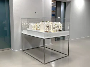 Architekturmodell auf weißem Sockel mit Acrylhaube, präsentiert auf einem metallischen Podest mit WINEGG-Logo in modernem Innenraum.