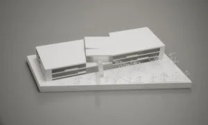 Weißmodell eines zweigeteilten Gebäudekomplexes mit Innenhof und abstrahierter Außenraumgestaltung.