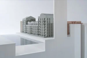 Wohnquartier-Modell mit grauen Bauvolumen und Innenhof, auf weißem Sockel mit Wasserfläche und Schnittdarstellung.