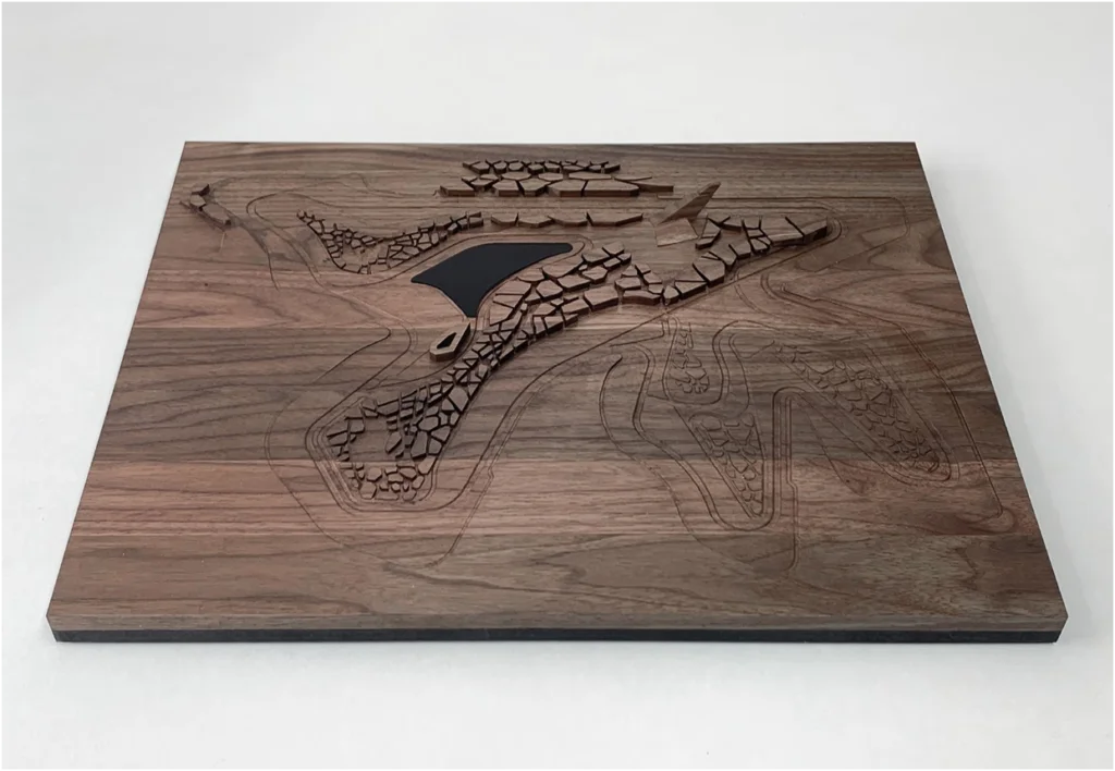Gesamtansicht eines topografischen Architekturmodells aus Holz mit gelaserten Geländelinien, fragmentierter Struktur und zentraler Wasserfläche