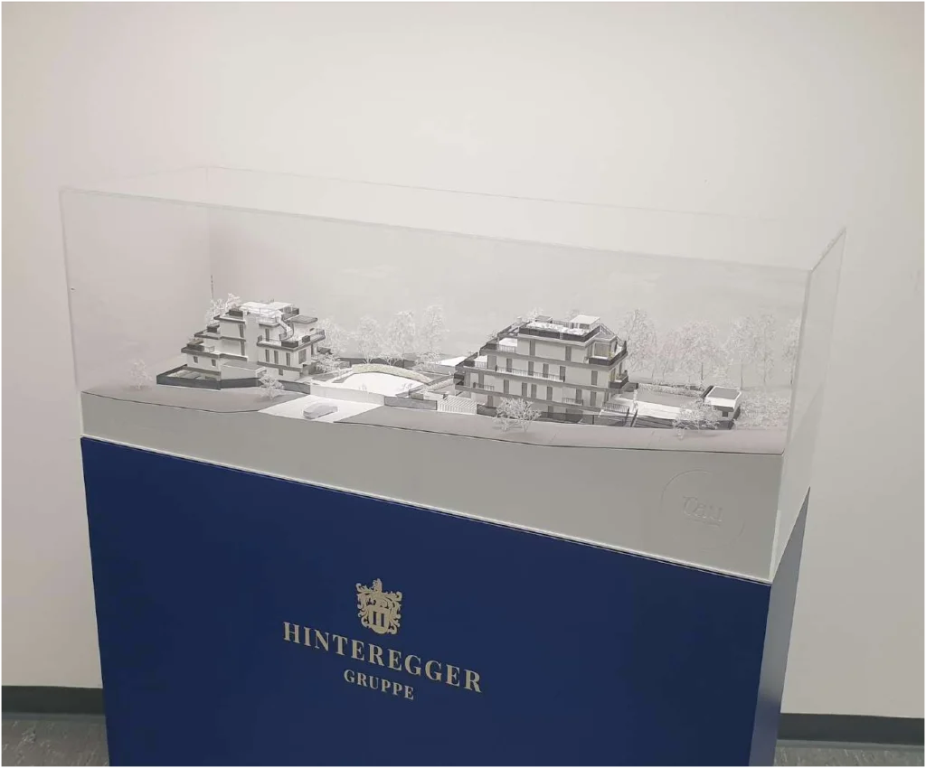 Individuelles Podest für das Modell – Sonderfarbe Blau mit Firmenlogo und Vor-Ort-Aufbau. Maßgefertigtes dunkelblaues Modellpodest aus folierten MDF-Platten mit silberner Logo-Prägung für eine exklusive Architekturpräsentation.